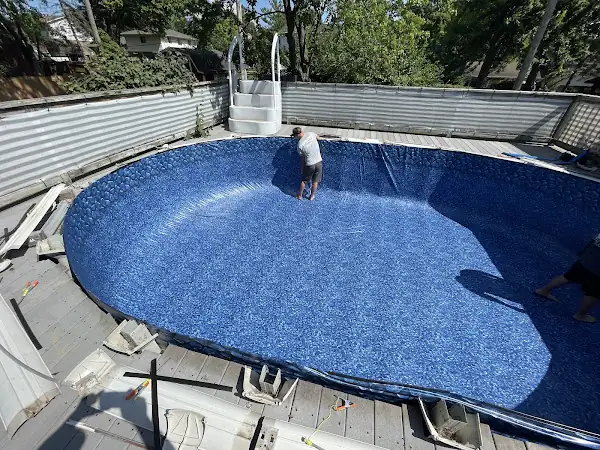 Two crew members inside an empty oval pool, laying a new blue liner with a stone border